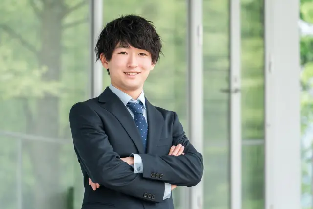 薄毛治療に前向きに取り組み、自信を取り戻した若いビジネスマンのイメージ写真。スーツ姿で腕を組み、笑顔を見せる20代男性が、AGAや若年性脱毛の早期対策の効果を体現している様子。薄毛改善後の前向きなライフスタイルを表現。