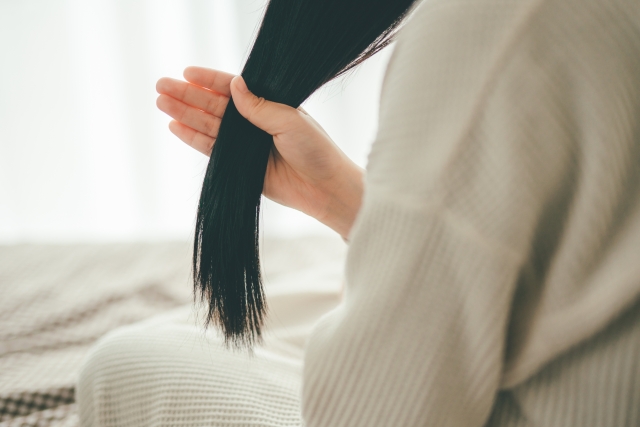 黒髪を手のひらと親指でつまみ、長く垂れた髪を見つめる女性の後ろ姿。髪質や色素の変化を意識する静かな瞬間|白髪と毛髪構造の科学的解説コラムより
