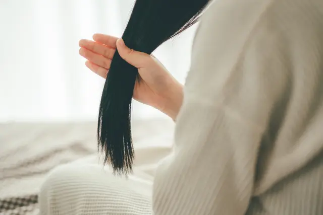 黒髪を手のひらと親指でつまみ、長く垂れた髪を見つめる女性の後ろ姿。髪質や色素の変化を意識する静かな瞬間|白髪と毛髪構造の科学的解説コラムより