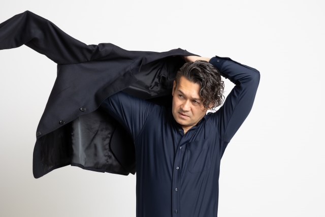 スーツを力強く羽織る日本人ビジネスマン。顔には無精ひげが残り、頭頂部の髪の毛がやや薄くなり始めているが、自信を持って身だしなみを整える姿が、若年性AGA（男性型脱毛症）と前向きに向き合う男性像を象徴している。