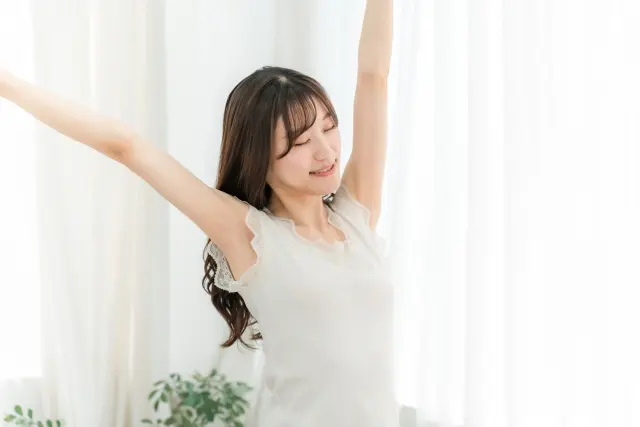 背中まで伸びたロングヘアの女性が両腕を伸ばしてストレッチする姿。頭頂部に軽度の薄毛が見られるが、通常は目立たない程度。女性にも適応される自毛植毛治療の可能性を示唆するイメージ
