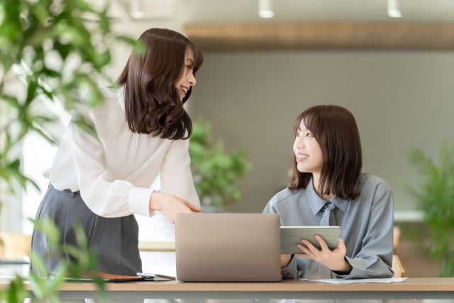 職場で談笑する2人の女性。ノートパソコンとタブレットを使って仕事中だが、どちらが自毛植毛を受けたかは全く分からない自然な髪型が印象的
