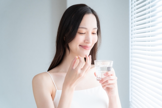 女性が薬を飲むために水と一緒に錠剤を手に取っている様子。窓から差し込む光の中で笑顔を見せており、スピロノラクトンやフィナステリドなど、女性型脱毛症（FPHL）の治療薬を前向きに服用している情景を象徴。髪の悩みに向き合いながらも希望を持って治療に取り組む姿勢を表現する本文と連動したイメージ。