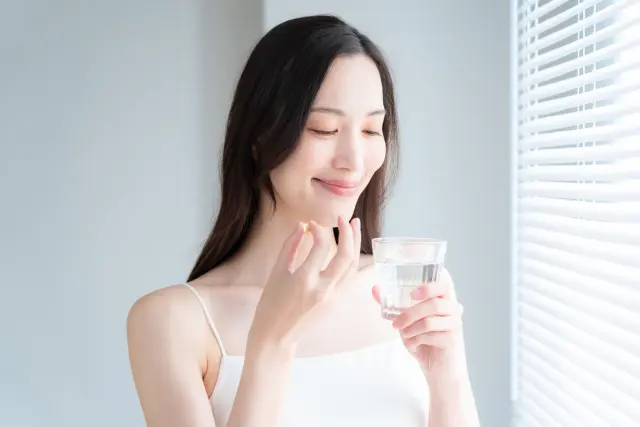 女性が薬を飲むために水と一緒に錠剤を手に取っている様子。窓から差し込む光の中で笑顔を見せており、スピロノラクトンやフィナステリドなど、女性型脱毛症(FPHL)の治療薬を前向きに服用している情景を象徴。髪の悩みに向き合いながらも希望を持って治療に取り組む姿勢を表現する本文と連動したイメージ。