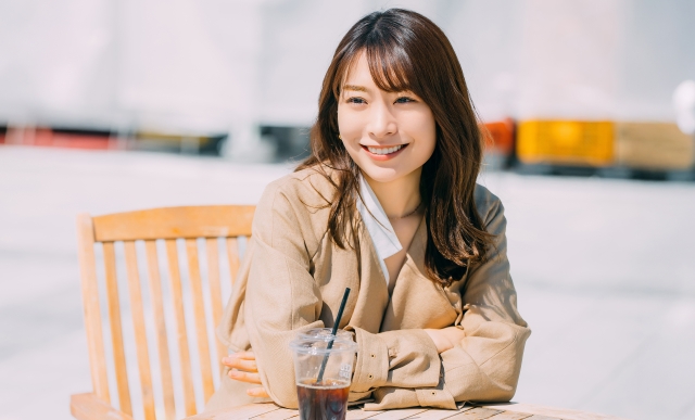 長いブラウンヘアの日本人女性がカフェのテーブルに身を乗り出し、太陽の光を受けてツヤのある髪を輝かせながら明るく笑っている様子。自然な毛流れと健康的な髪質が際立ち、毛髪移植後の理想的な仕上がりをイメージさせるビジュアルカット。