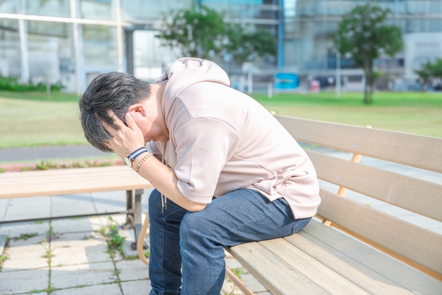 薄毛の進行に悩み、公園のベンチでこめかみに手を当てて思い悩む50代の男性。髪のボリューム減少やフィナステリドの副作用への不安など、脱毛症に対する心理的ストレスを表現したイメージ写真。