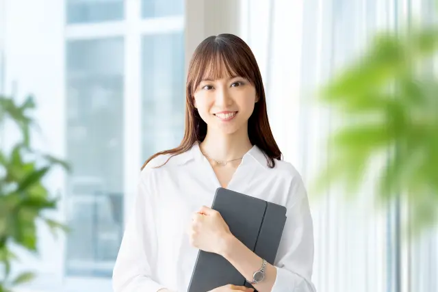 白いブラウスを着た女性がタブレットを胸に抱えながらカメラに向かって明るく微笑む姿。シルクのような艶のある髪が肩から背中にかけて美しく流れ、健康的な髪の印象が女性の魅力と自信を引き立てている。