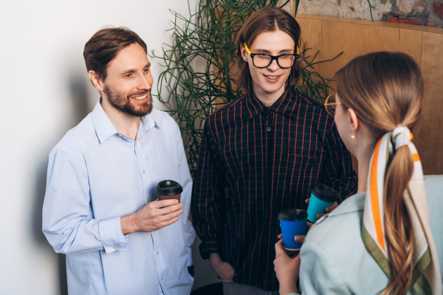 ヒゲをたくわえた男性が女性同僚2人とオフィスでコーヒーを片手に立ち話をする様子。ヒゲが印象的な男性の姿が、フェイシャルヘアスタイルによる見た目の変化と自信の向上を象徴している。ヒゲ移植後の自然な印象を伝えるイメージカット。