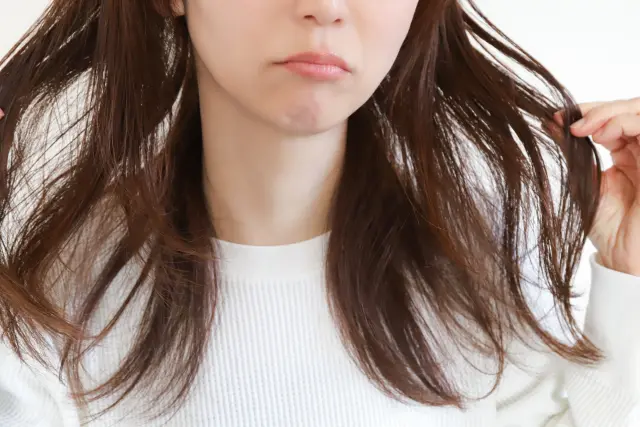 口元に不安げな表情を浮かべた女性が、両手で広がった長い髪をつまんでいる様子｜抜け毛や薄毛の悩みを抱える女性の心理的ストレスを象徴するイメージ｜女性の脱毛症コラム用装飾画像