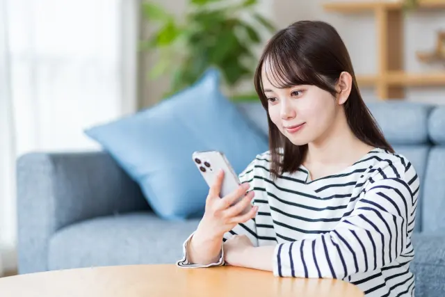 ストライプ柄のシャツを着た若い女性がローテーブルに座り、スマホで髪の悩みに関する記事を読む日常のワンシーン