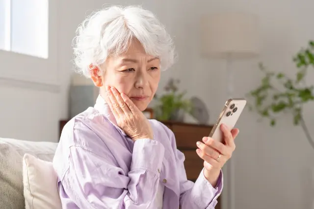 白髪の高齢女性が鏡を見つめながら頬に手を添えてため息をついている様子。年齢や髪色による見た目の変化に悩む女性の気持ちを象徴し、高齢者でも可能な植毛治療の必要性や関心を表現したイメージ。