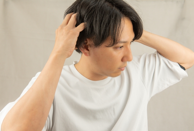 若い男性が髪の中に指を差し入れ、地肌が露出している部分を探るように頭皮を触っている。表情は無表情ながらも集中している様子がうかがえる。