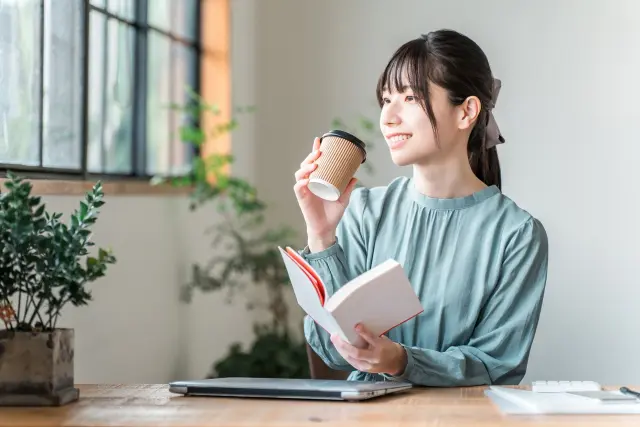 窓際の明るい木製テーブルでコーヒーを片手に読書を楽しむ笑顔の女性|コーヒー由来成分の美容・健康効果を紹介するヒロクリニックのライフスタイルコラム用イメージ
