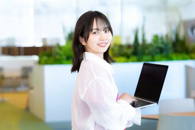職場でノートパソコンを抱えながら微笑む20代の女性。髪のボリュームや質感が気になり始める世代のイメージカット