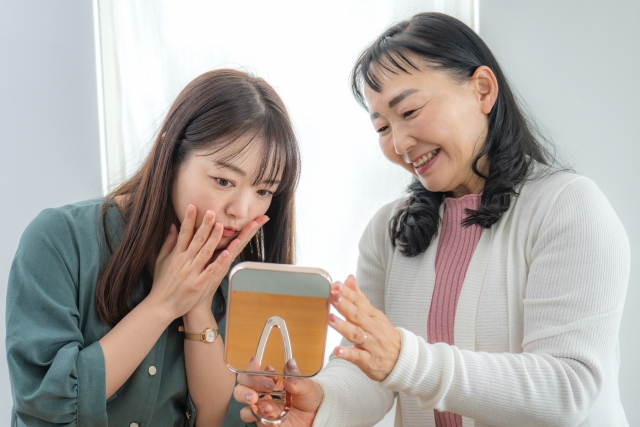 手鏡を見ながら薄毛の悩みを語り合う高齢女性と若年女性のイメージ。世代を超えて進行する女性の抜け毛やボリュームダウンの不安を象徴する場面。