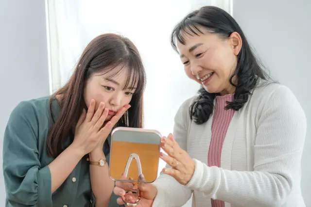 手鏡を見ながら薄毛の悩みを語り合う高齢女性と若年女性のイメージ。世代を超えて進行する女性の抜け毛やボリュームダウンの不安を象徴する場面。