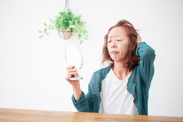 鏡を見つめて薄毛に悩む高齢女性と、生き生きと茂る観葉植物の対比を通じて、加齢による女性の薄毛や脱毛症の悩みを表現したイメージ｜脱毛縮小術による治療検討の文脈で使用