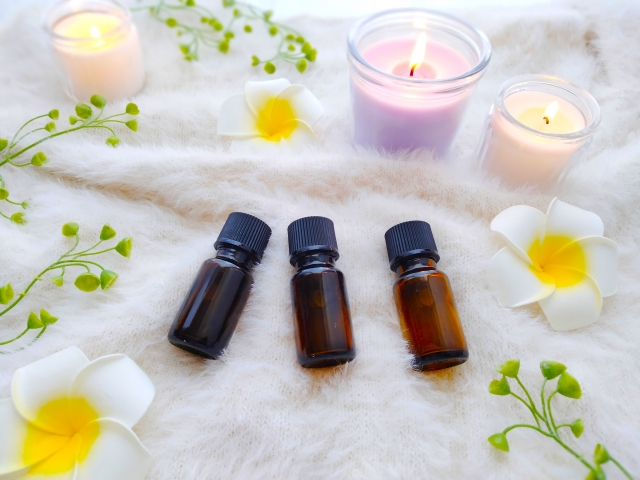 白いウールラグの上に並べられたエッセンシャルオイルのガラス瓶と、点灯されたパステルカラーのキャンドル、観葉植物風の装飾。自然派ヘアケアや精油を用いたリラックス空間の演出をイメージさせる装飾用写真。