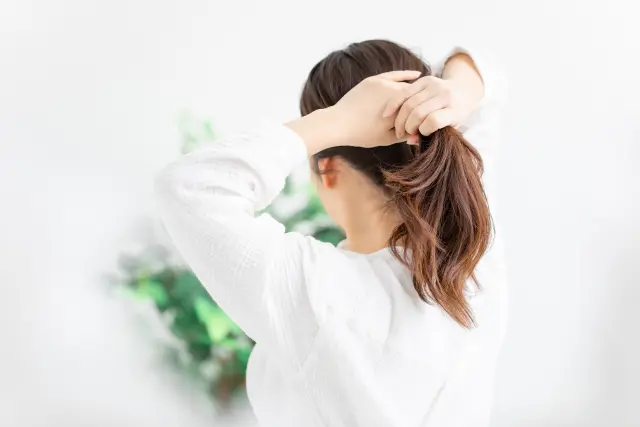日差しが差し込む窓辺で、両手を使って髪を束ねる女性。白いシャツに光が反射し、黒髪とのコントラストが印象的。ポニーテールにする瞬間、頭頂部や分け目の地肌が自然に露出し、女性特有の薄毛のサインが浮かび上がる。