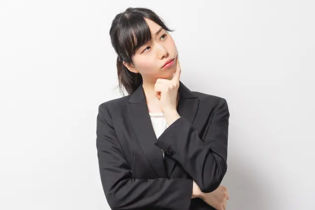 前髪の分け目がやや目立つ女性ビジネスパーソンが、頬に指を当てて物思いにふける様子。仕事やストレスによる抜け毛、女性の薄毛の悩みを連想させるビジュアル。女性型脱毛症や頭皮ケアへの関心を高める医療記事の装飾画像に最適