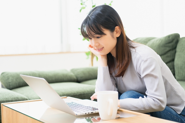 自宅で薄毛ケアの情報を調べる若い女性が、ソファに座って笑顔でパソコンを操作している様子｜女性のための薄毛治療・自宅ケア特集コラム用イメージ