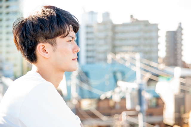 高層ビルの屋上で街を見下ろす若年男性。強い日差しが頭頂部を照らし、髪の薄さをほのめかす。将来の脱毛への不安や自信喪失を象徴する、憂いを帯びた表情の後ろ姿。