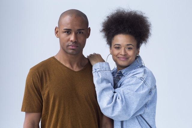 自然な縮毛（ナチュラルヘア）を上向きに立ち上げたアフリカ系女性と、その隣で寄り添うパートナー。髪型を通じた文化的アイデンティティの表現や、ストレートヘア信仰からの解放、多様性の受容を象徴するビジュアル。牽引性脱毛症のリスクを避けた自然なヘアスタイルの肯定的事例としても文脈に適合。