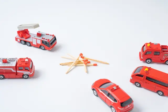 おもちゃの消防車がマッチの山に向かって並ぶイメージ。火災事故による外傷や重度熱傷が原因で頭皮に瘢痕が残り、脱毛に悩む人々の存在を象徴的に表現。無償植毛支援プログラム「オペレーション・リストア」に関連するビジュアル。
