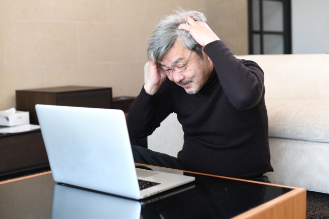 ノートパソコンの前で頭を抱える白髪の高齢男性｜海外植毛手術の失敗や費用トラブルに直面した後悔や不安を表現したストック写真