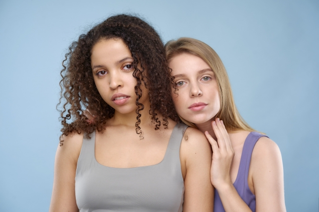 異なる人種の女性二人（アフリカ系と白人系）が並んでポーズをとる姿。髪型や美しさの多様性、文化的背景の違いを象徴するビジュアルで、固定的な美の基準への問いかけやヘアアイデンティティの尊重を訴える文脈に適した画像。