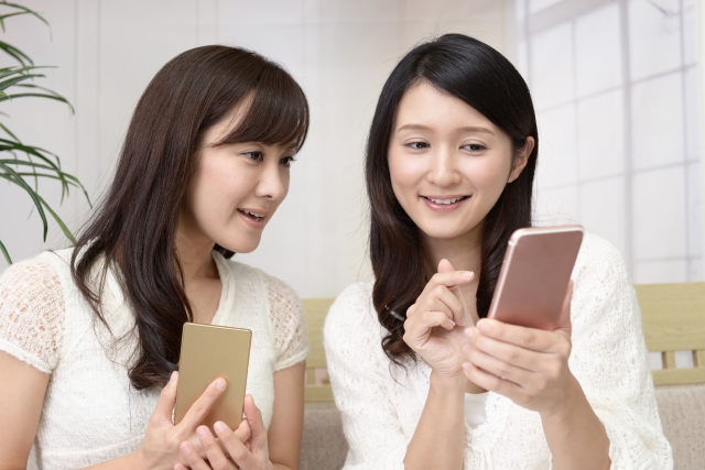 スマートフォンの画面を見せながら会話する主婦たち。育毛剤の口コミやビフォーアフター画像などを共有しながら情報交換する様子は、証言ベースの広告に影響を受けやすい現代女性の傾向を象徴。