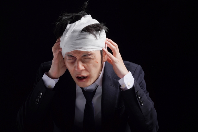 スーツ姿の男性が頭に包帯を巻き、脱毛や外傷による痛みに苦しむ様子。やけどや事故による脱毛に悩む人々が無償植毛手術で希望を見出す「オペレーション・リストア」プログラムの文脈を示すイメージ。