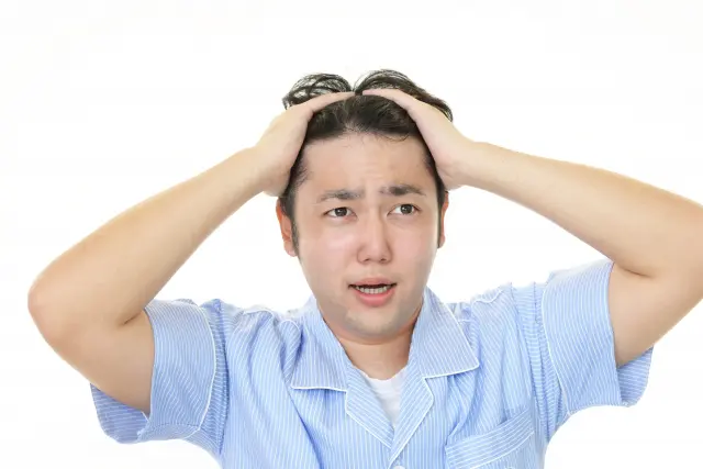 前髪の生え際が後退し始めたことにショックを受け、両手で髪をかき上げながら額を見つめる若い男性。進行する薄毛やAGAに対する若年層の不安を表現したイメージ。
