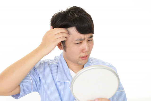 生え際の後退に気づき、不安そうに鏡を見る若い男性の様子。20代で自毛植毛を検討する前の悩みや焦りを象徴するシーン｜若年性AGAの進行と外見への不安を表現