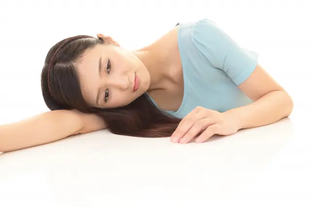 机にうつ伏せてうなだれる中年の日本人女性。一見すると髪は長く艶やかだが、前頭部から頭頂部にかけて地肌が透けるようなびまん性脱毛が目立ち始めている。薄毛の悩みを抱え、孤独や不安を感じている様子。