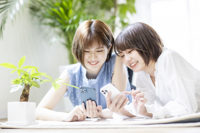 明るい部屋で寄り添いながらスマートフォンを見て笑い合う女性たち。SNS広告や口コミで育毛情報を共有し合う、現代女性のリアルな日常シーンを表現したイメージ。