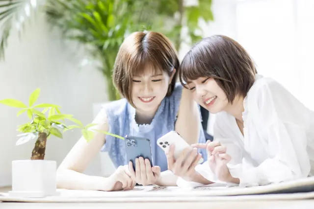 明るい部屋で寄り添いながらスマートフォンを見て笑い合う女性たち。SNS広告や口コミで育毛情報を共有し合う、現代女性のリアルな日常シーンを表現したイメージ。